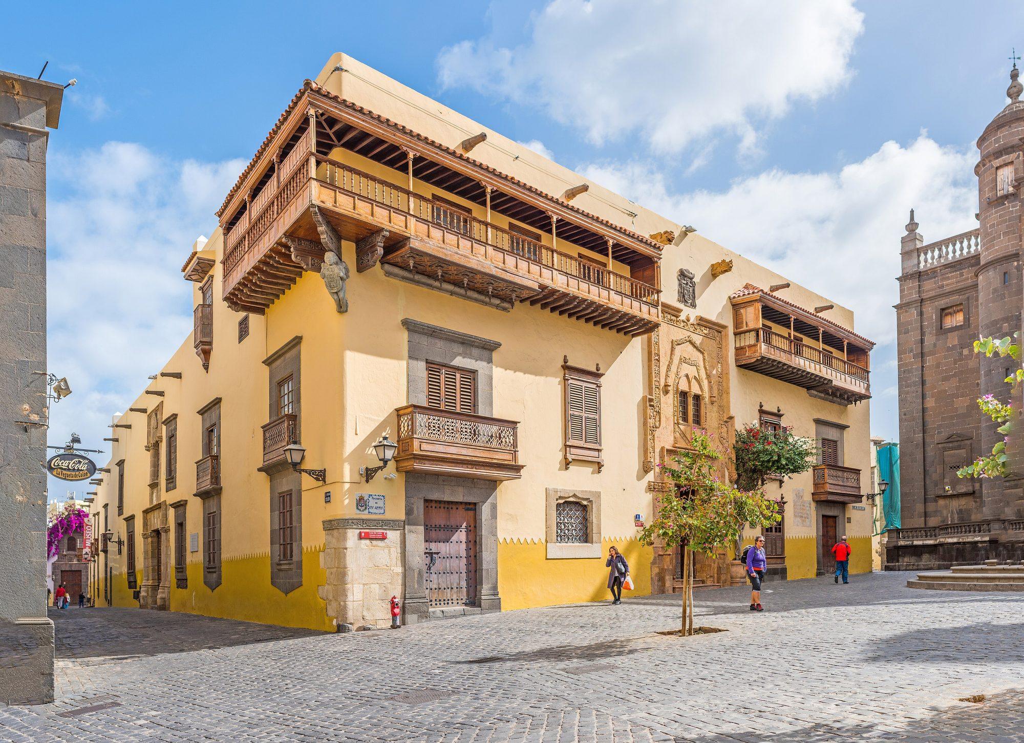 The corner of the Casa de Colón in Vegueta, Las Palmas: an ochre lime-washed house with a long carved wooden gallery balcony under a tiled roof, smaller wooden balconies with lattice fronts above windows and doors framed in dark basalt, and a carved stone portal, on a cobbled street that runs off to the left, with the dark stone flank of the Catedral de Santa Ana on the right, under a blue sky with white clouds