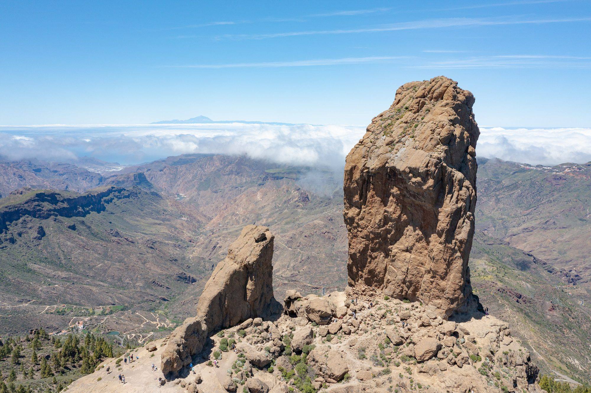Roque Nublo, a tall rock monolith, and the smaller El Fraile on a rocky summit plateau in the centre of Gran Canaria, with hikers at their foot, above pine-dotted ridges and deep ravines, a sea of clouds beyond and the peak of Teide on Tenerife on the horizon