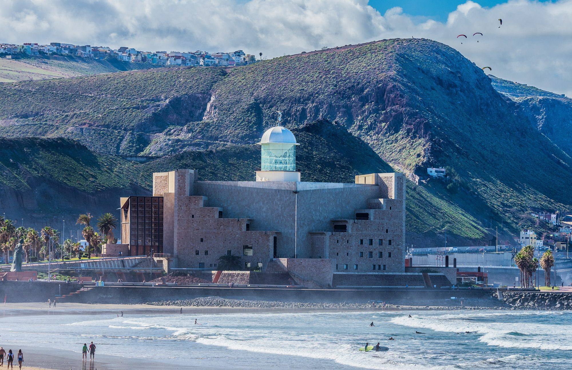 The Auditorio Alfredo Kraus, a stone-clad block crowned by a glass lantern under a white dome, at the end of Las Canteras beach, with surfers in the breaking waves and walkers on the sand, green hills with colourful houses along the ridge behind and paragliders in a cloudy sky