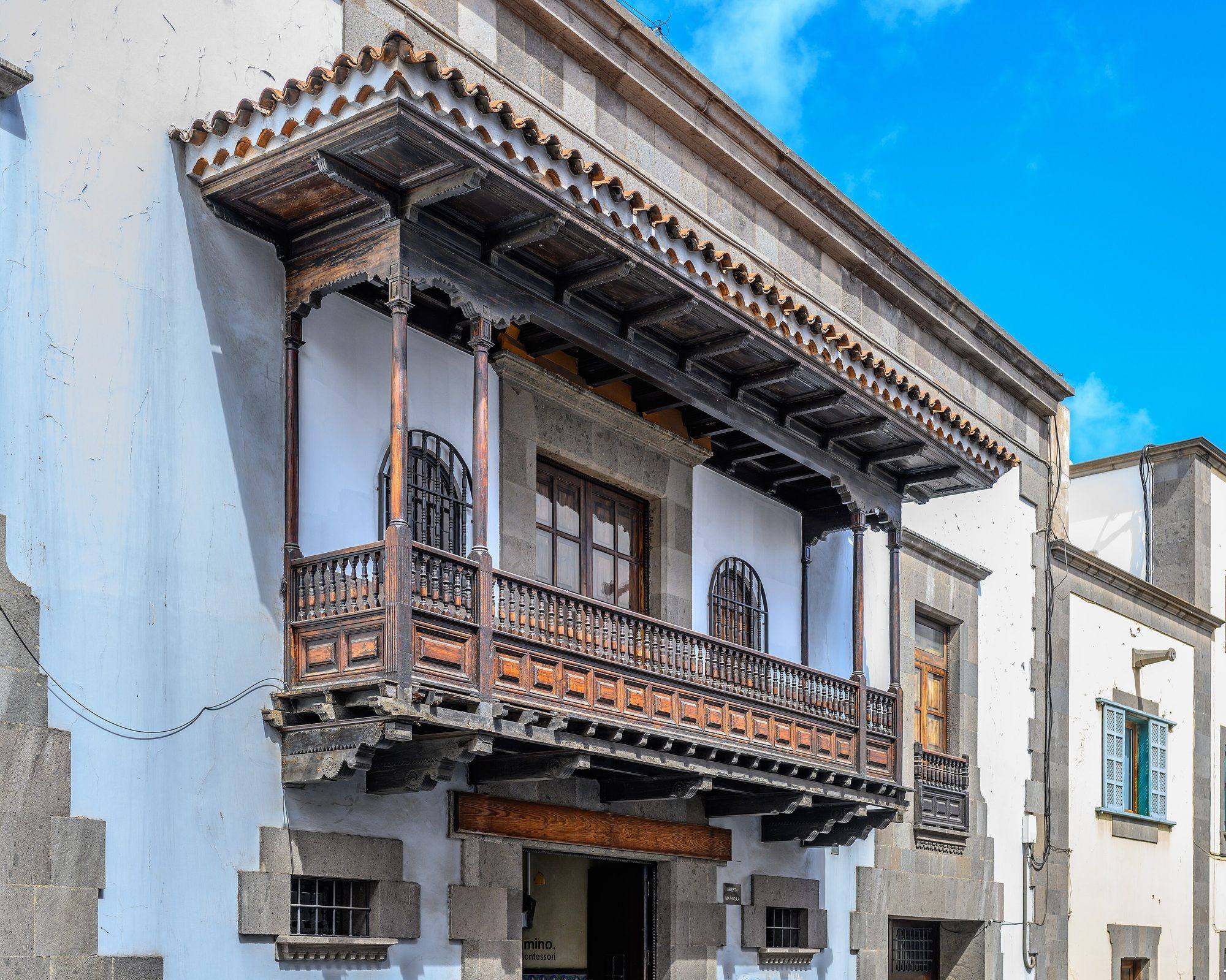 A long Canarian wooden balcony on a white lime-washed house in Las Palmas: slender dark wooden posts holding a deep roof edged with clay tiles, a railing of turned balusters above panelled sides, the whole resting on carved wooden corbels, set against grey basalt quoins and window frames, with a pale blue latticed shutter on the neighbouring house, under a bright blue sky
