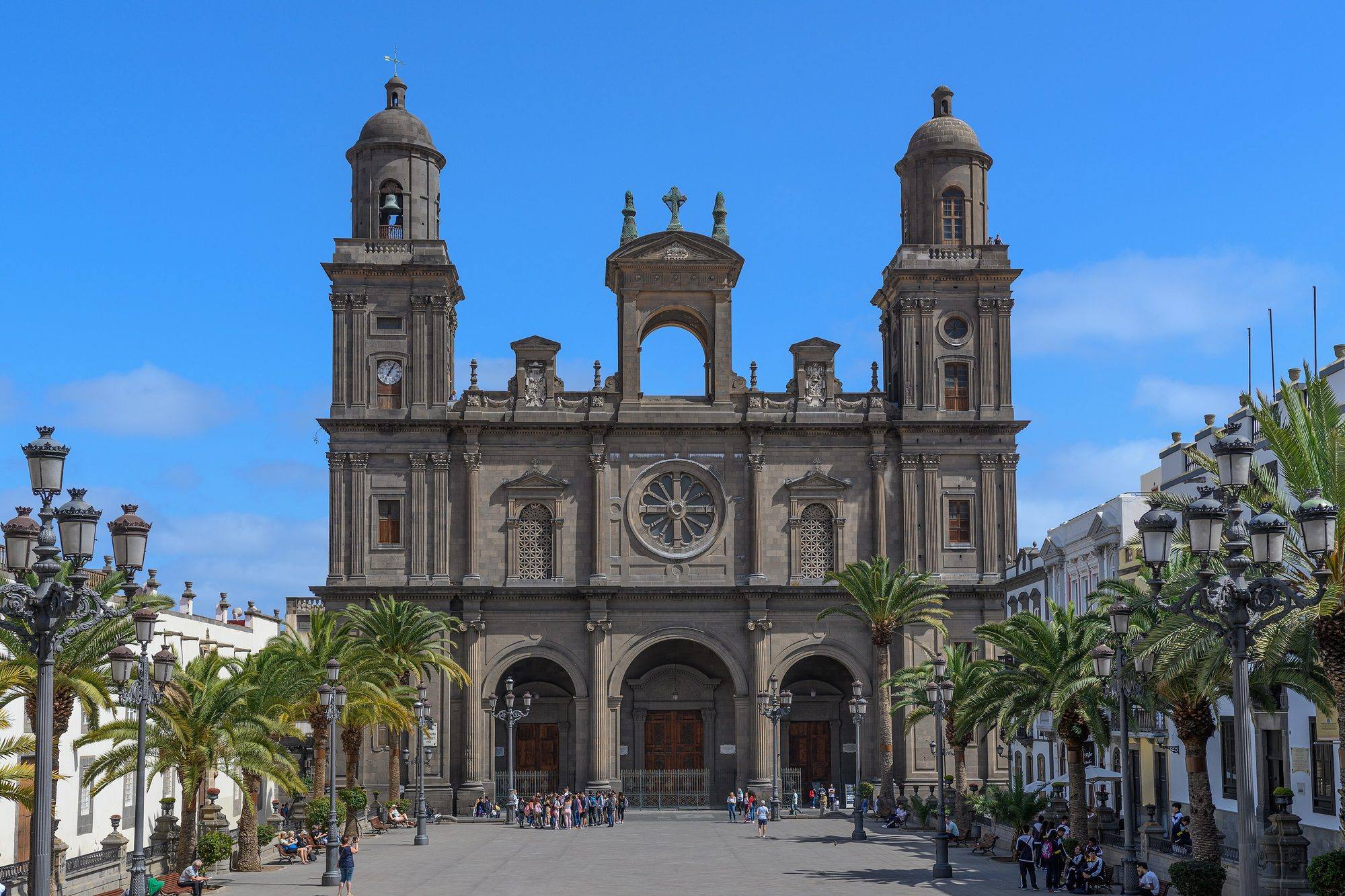 The main front of the Catedral de Santa Ana in Vegueta, built of dark grey stone, with two domed bell towers, a central open arch topped by a cross, a rose window and three arched doorways, at the end of the Plaza de Santa Ana lined with palm trees, ornate iron lamp posts and white houses, under a deep blue sky